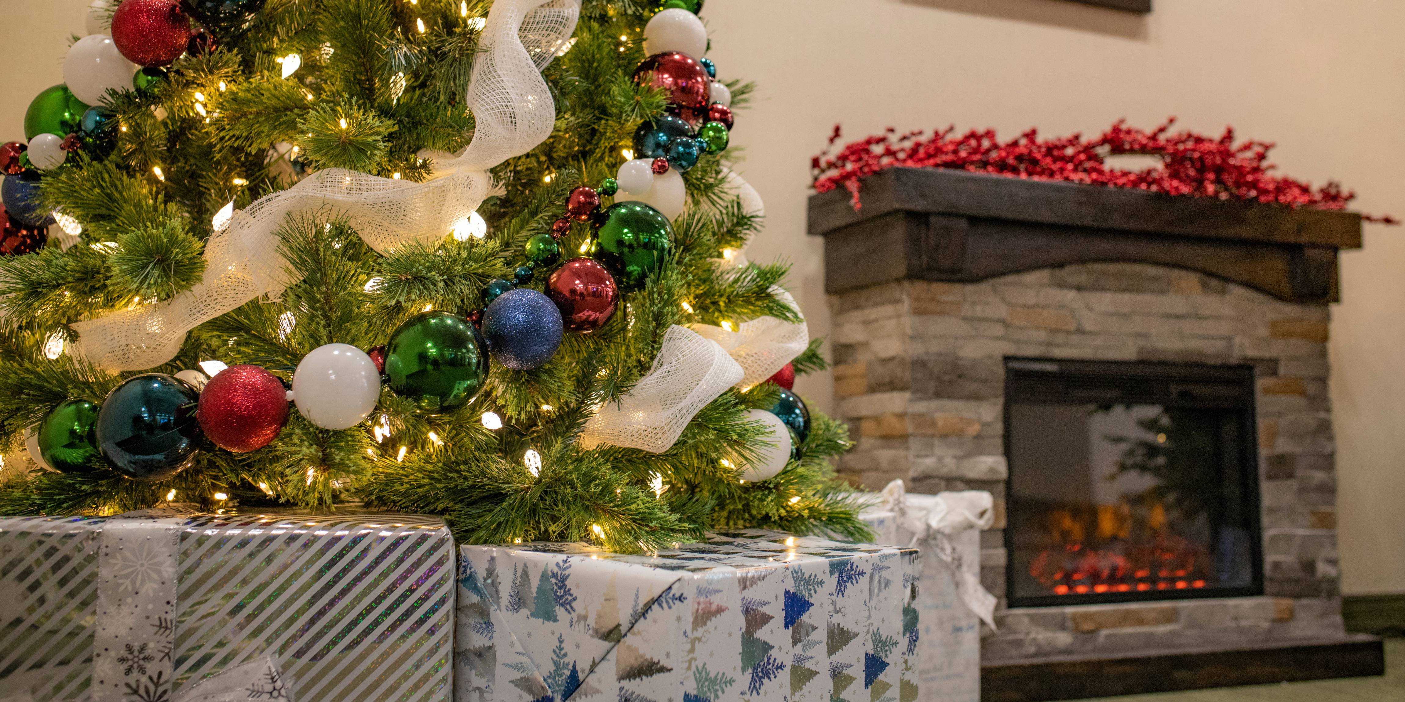 Green artificial christmas tree with red, blue, and pink ornaments and white garland. Presents are around the tree trunk and a fireplace roars fire in the background.