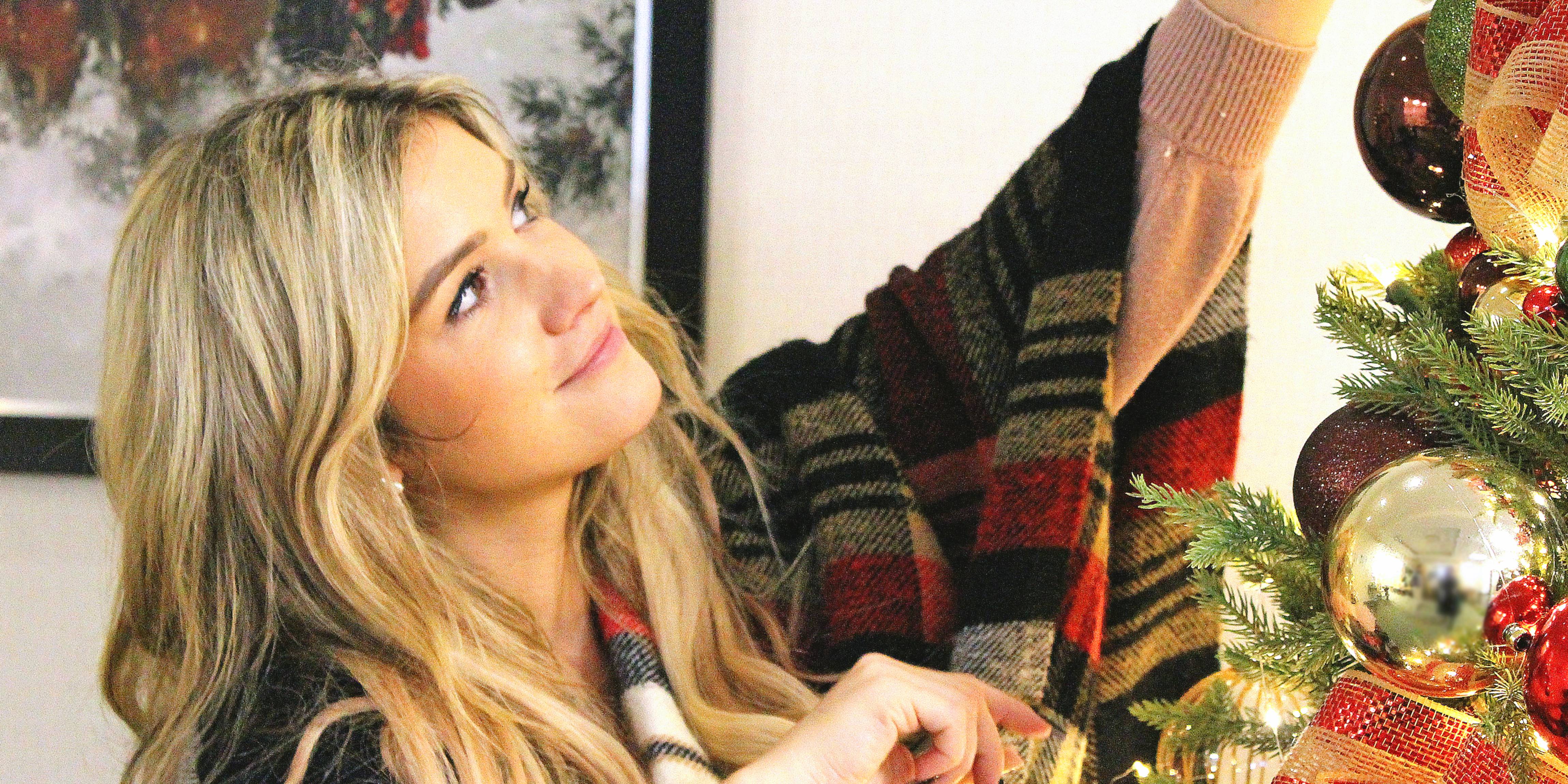 Blonde girl with checkered blanket decorates an artificial green christmas tree. The tree has gold, brown, and red ornaments as well as red ribbons.