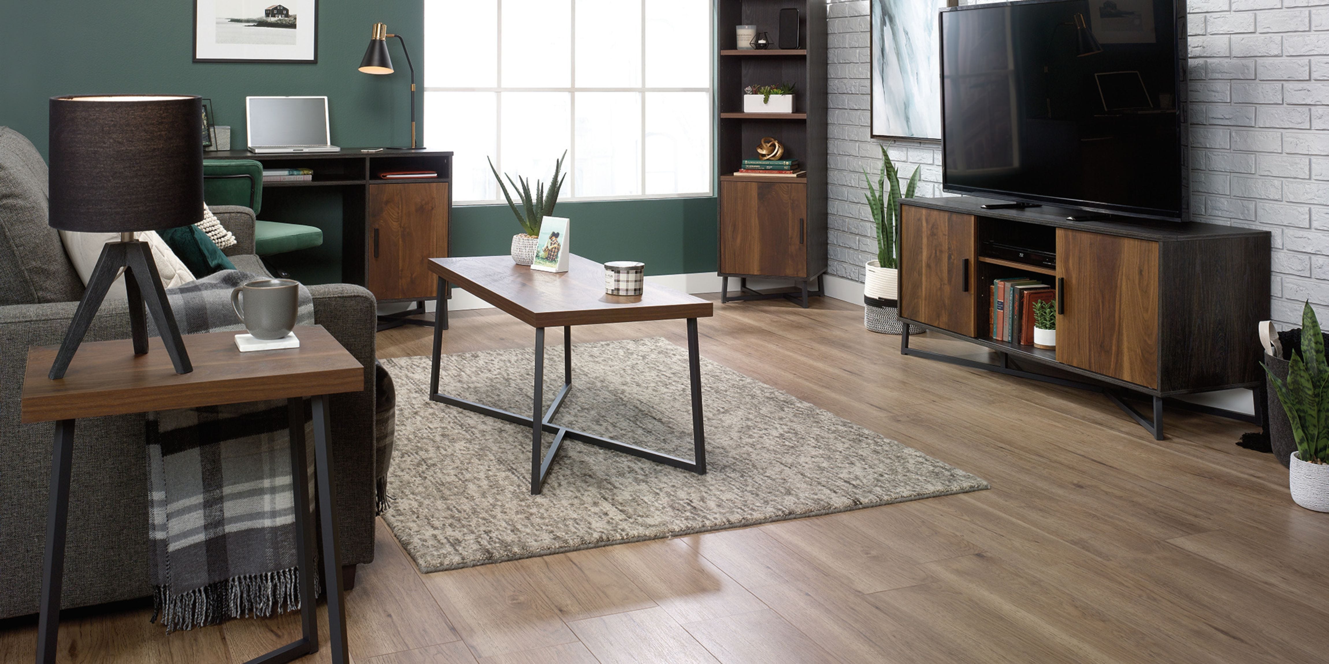 Large living room with one green wall and one white brick wall. On the right, a wooden TV stand with a flat screen TV. On the left, a grey couch with a wooden side table with a lamp on top. A beige carpet can be seen with a wooden coffee table and a plant