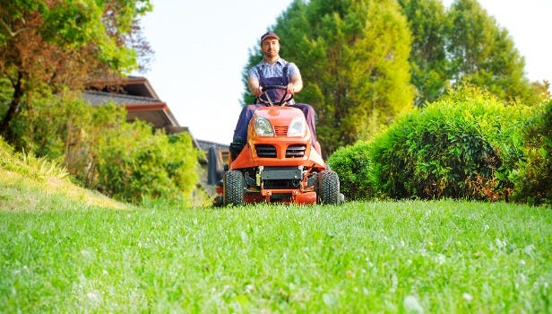 Man with ball cap on bright orange tractor mowing green grass. Trees and bushes in the background.