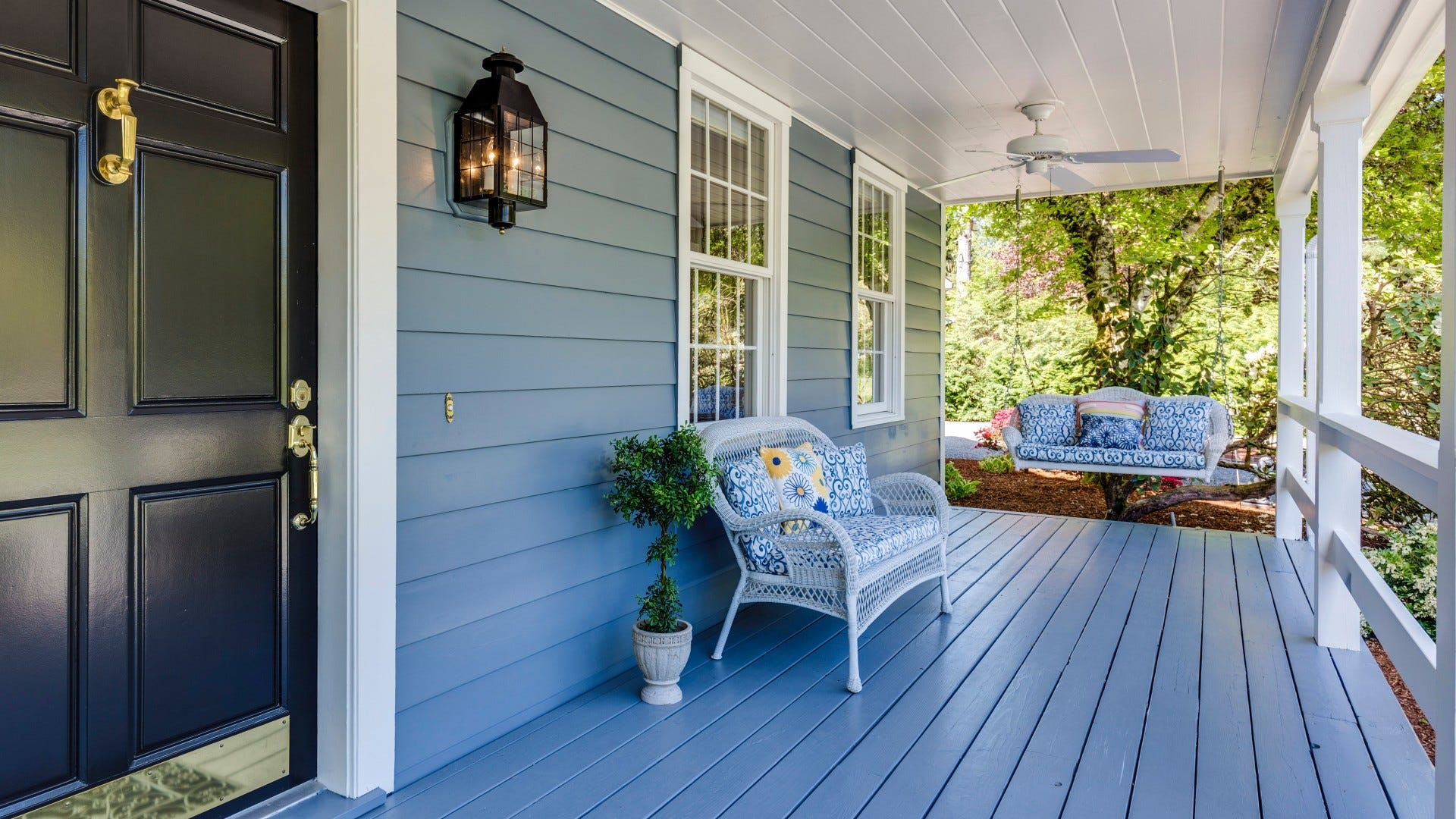 Blue sided home and deck. Black door next to two large windows. Chair with accent pillows sit under windows. Hanging chair with accent pillows hangs in the back part of the deck.