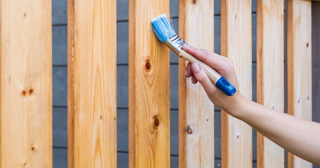 Hand coming from the right side of the screen holding a medium beige paintbrush with blue bristles and a blue tip. The brush is staining the side panels of a deck. The first 3 are a dark brown stain color and the last 4 are lighter and not yet stained.